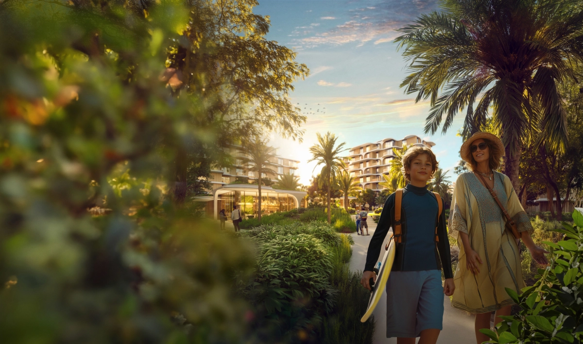 Family walking with surfboard at The Beach House Fahid, surrounded by lush greenery and modern buildings.